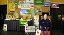 Photo of a booth display at an event, with various posters and other informational materials displayed, and a person seated next to the table.