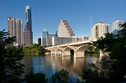 Photo of the Austin city skyline on a sunny day, with an assortment of large downtown buildings, varying in style and shape, in the background and a river and bridge in the foreground.