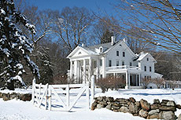Photo of a snowy residential scene, with a large, ornate house on a hill in the background, with trees beside it, and a fence and rocky border in the foreground.