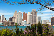 Photo of the Detroit city skyline on a sunny day, with a variety of office buildings clustered together and the Detroit River flowing beside.