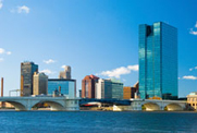 Photo of a city skyline awash in blues, with a river and bridge in the foreground and several large buildings behind, against a brilliant, clear sky.