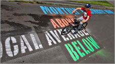 A photo of a bicyclist riding along a paved surface with painted lettering on it.
