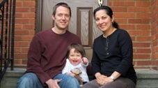 A photo of a family, with father, mother, and toddler in the middle, sitting on the steps in front of a brick home.