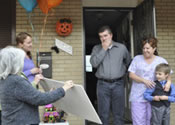 Photo of a family -- father, mother, and child -- standing at the doorway of a brick home, facing a group of people holding a sign up to them.