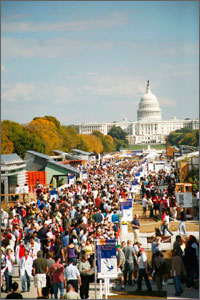 Photo of a colorful crowd of people walking among modern-looking houses that line two sides of a street. In the background, trees are changing from green to orange and the U.S. capitol shines white against a blue sky.