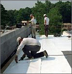 Photo of a man kneeling on one knee and applying a white material to a black roof. Three other men are standing in the background with their backs to the camera. 