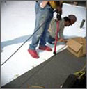 Photo of two workers on a roof installing light-colored roofing material. 