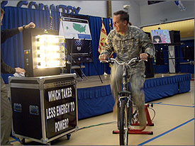 Photo of a man in uniform riding a stationary bicycle and looking at an illuminated light bulb display.