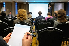 Photo of people watching a presentation on a screen; the foreground shows a person's hands taking notes on a notepad.