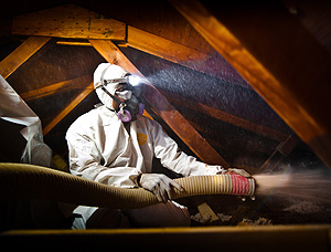 This photo shows a man in a white hazardous materials suit blowing insulation inside of an attic. He is wearing a headlamp on his head and the beam shines in the general direction of the insulation tube he is holding.