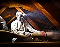 This photo shows a man in a white hazardous materials suit blowing insulation inside of an attic. He is wearing a headlamp on his head and the beam shines in the general direction of the insulation tube he is holding.