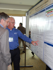 Photo of two men standing in front of a mounted poster. One of the men is pointing at the poster.