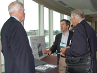 Photo of three men talking in front of a table. A small sign and a light fixture are on the table.