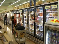 Photo of a row of freezer cases at a supermarket with a ladder and workcart nearby and a maintenance worker standing next to them.