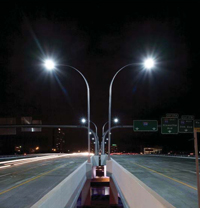 View at night looking down a bridge made up of two sections. Between the sections is a base holding two bright street lamps, shaped liked antennas, the left one curved to the left and the right one curved to the right.