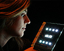 Photo of a woman's head in a dark room looking at a square object containing two horizontal rows of bright white circles of light, with a row of what appears to be white text in the middle.