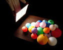 Photo of a rectangular white light panel containing grid lines being held by a person on the left (you only see one of the person's hands), shining the light onto a table to the right containing multi-colored balls.