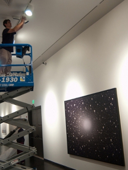 A man stands on a small crane installing a light fixture to illuminate artwork on a wall.