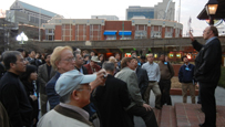 Photo of a large group of people outside listening to a man speak.