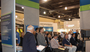 Photo of several LIGHTFAIR attendees sitting and standing inside the DOE SSL booth as they view an exhibit presentation.