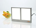 Photo of a horizontal rectangular device containing two white vertical panes side by side within a silver frame, with a cord coming out the back, sitting on a shiny surface, with a flower on its left.