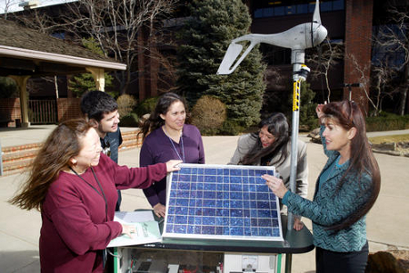 Photograph of a group of young women gathered around a wind power experiment, with a building and a wooded area in the background.