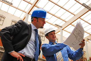 Two men in an industrial environment wearing hardhats examining a blueprint