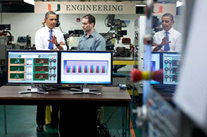 Photo of President Obama talking with another man and motioning towards two computer screens showing graphs.