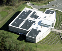 Photo of a white industrial building with a white roof featuring photovoltaic panels on top. The building stands on a verdant lot of land with a parking lot to the right.