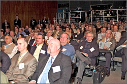 A large crowd of people, some in rows of chairs and some on bleachers, sit attentively while holding notebooks and agendas.