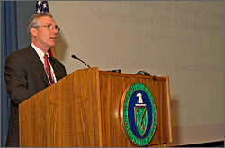 Steven Chuslo, from the U.S. Department of Energy, stands behind a podium in front of a large projection screen. Information about the TEAM Initiative and DOE's Renewable Energy Forum is displayed on the screen.