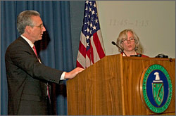 Steven Chuslo and Anne Crawley, from the U.S. Department of Energy, stand behind a podium in front of a large projection screen and a large U.S. flag.