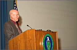 Scott Richlen, Acting FEMP Program Manager, stands behind a podium in front of a large projection screen. Information about the TEAM Initiative and DOE's Renewable Energy Forum are faintly visible on the screen.