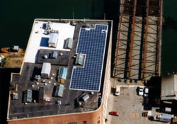 Aerial photo of a building with an area of the roof covered in PV panels, arranged tightly around other structures that from the air appear to be small sheds and access structures.