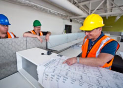 Photo of three men standing together reviewing a large print document. The men have bright orange and yellow construction vests and hard hats.