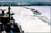 Photo of pipes floating on the Pacific Ocean with a barge floating alongside. Several people are photographed standing on the barge.