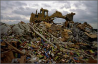 Photo of a bulldozer on top of a large trash mound in a landfill with a cloudy sky in the backdrop.