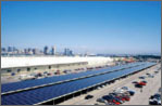 Photo of a long PV panels on top of a parking garage in a suburban area. Cars are also pictured next to the PV panels.