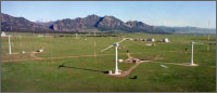 Photo of multiple wind turbines stand on green space in front of a mountain backdrop.