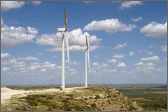 Photo of two wind turbines standing on a mountain in front of a cloudy blue sky.