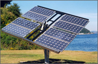 Photo of a square, tracking, standalone photovoltaic array stands in front of a tree and body of water.