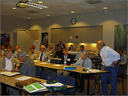 This photo shows workshop participants meeting in a conference room. Some of the participants are seated at tables looking at documents, while others are standing.