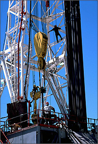 Photo of a well drilling rig. A man stands on the platform while another is climbing the rig.
