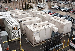 Photo of seven fuel cells with a series of pipes to the side in a parking lot setting. Steam is rising above some of the fuel cells.