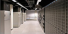 Photograph showing a view down a long hallway toward a wall. On the right hand side of the hallway is a vertical metal structure with a grid-like pattern on the structure. On the left hand side is a row of refrigerators. Lights run the length of the ceiling.