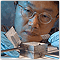 Photo of a man in a lab coat, wearing glasses, who is working on a small, metallic, square device.