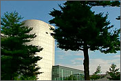 Photo of a building with a large, white cylindrical tower next to a small, squat building with large glass windows covering its surface. The building is mostly hidden behind trees. Photo links to the Federal Energy Management Program, National Institutes of Heath Showcases Utility Energy Services Contracts.