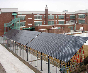 Photo of an array of solar panels mounted to poles that attach to the ground. A school is located in the background.