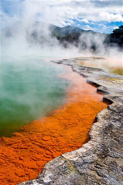 Photo of steam rising above a pool of water. A range of mountains and sky rise in the background.