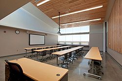 Photo of a daylit classroom that has four long wooden tables and chairs, a teacher's desk, projector, and projector screen. The classroom has reclaimed Douglas fir wood paneling along one wall and the ceiling, and polished concrete floor.